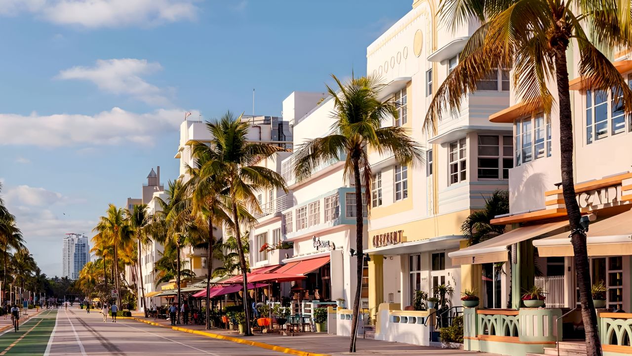 A wide view of a street with buildings, palm trees, and people walking.