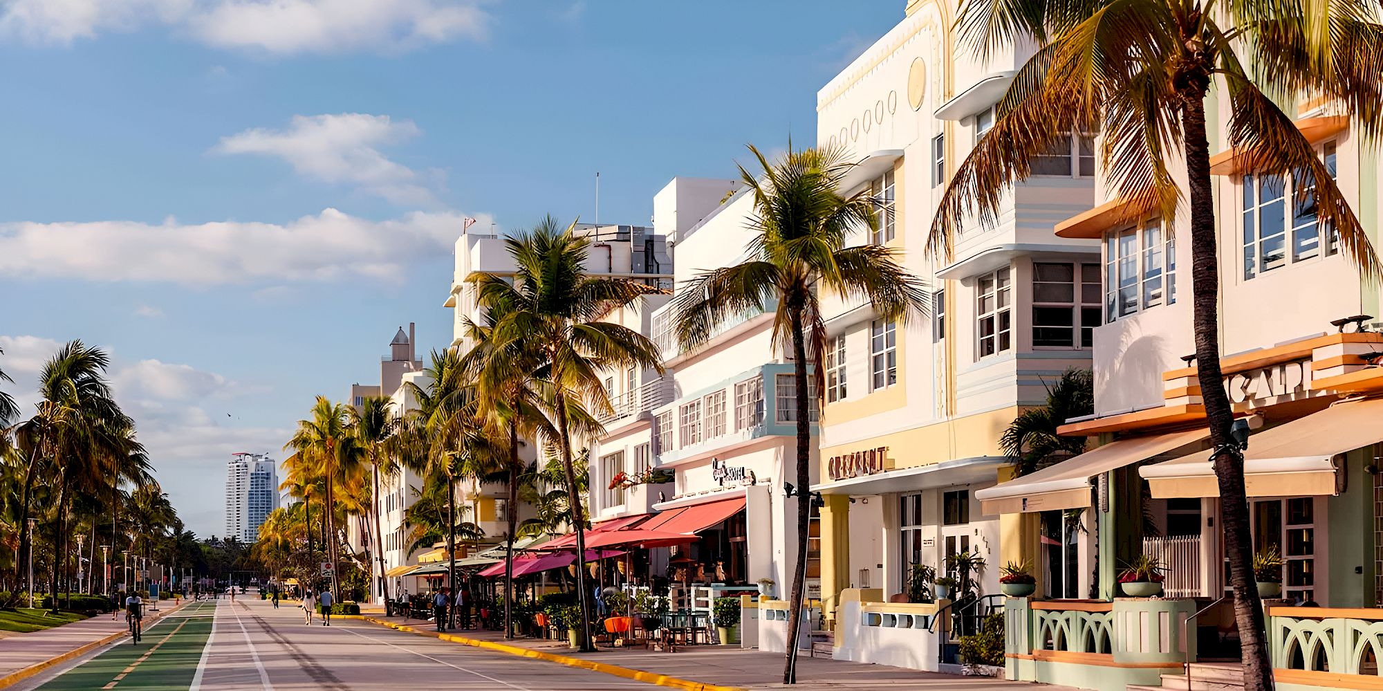 A sunny beachfront street with palm trees, pastel storefronts, and outdoor caf&eacute;s along a paved promenade.