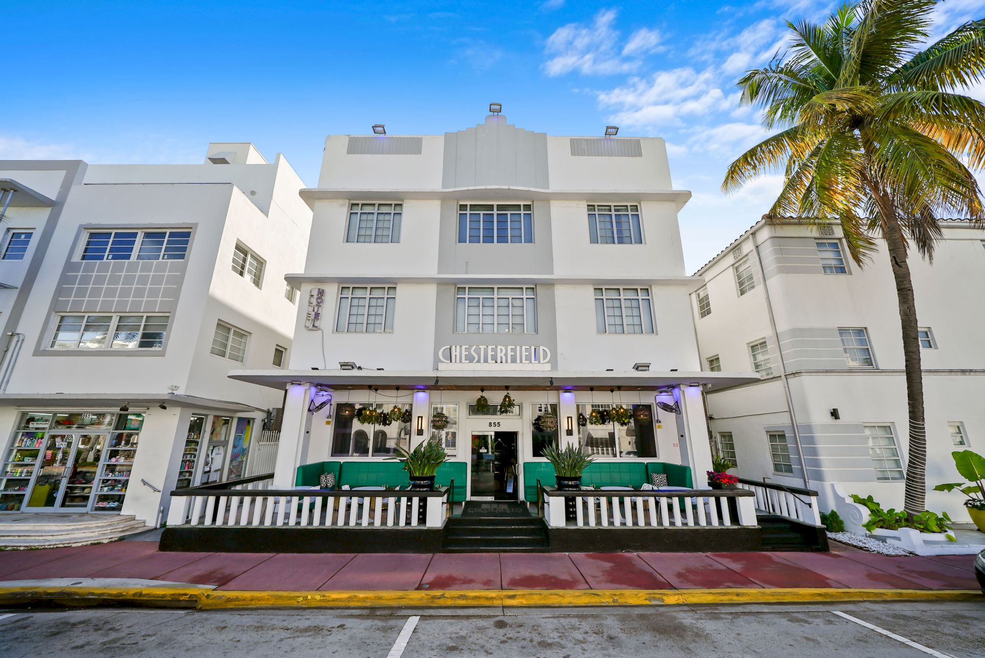A white Art Deco-style hotel with purple signage and a shaded porch, turquoise seating, and a tall palm tree on the right. End.