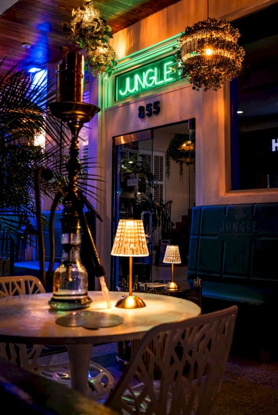 A cozy cafe scene with a round table lit by a small lamp, lush plants, and a neon &ldquo;Jungle&rdquo; sign outside the doorway, creating a warm, tropical vibe.