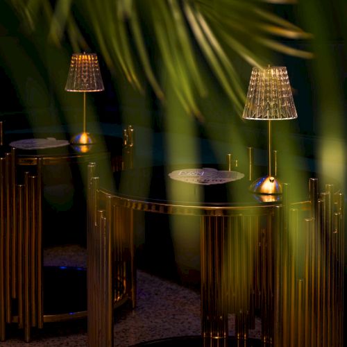 Dimly lit lounge with gold-accented chairs and hanging lamps, seen through plant fronds, inviting intimate nighttime seating.