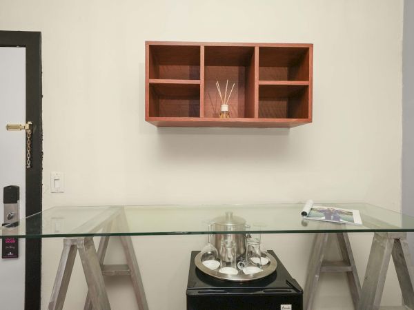 A minimalist office setup with a glass desk, a small wall shelf above, and a few items on the desk and shelf.