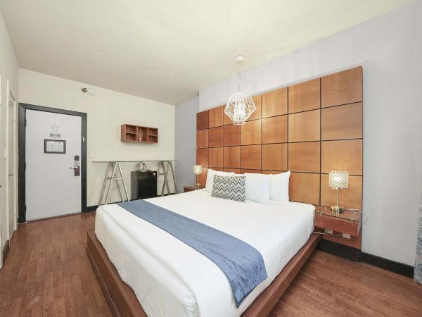 A modern hotel bedroom with a large bed, wooden paneled headboard, two nightstands, a hanging lamp, and a small desk near the door.
