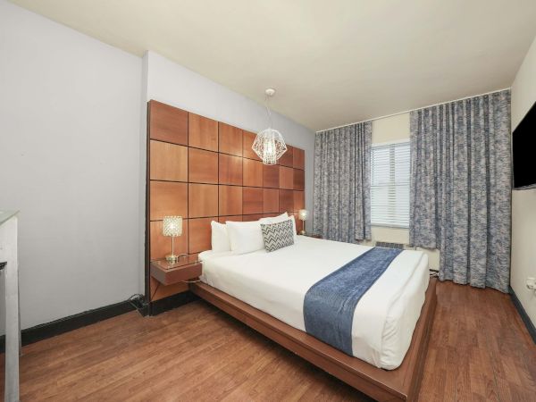 A modern hotel room with a large bed, a wooden headboard, blue accents, bedside lamps, a small desk, and a window with patterned curtains, nice and neat.