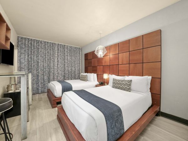 A modern hotel room with two neatly made double beds, a padded brown headboard, a small desk and chair, a flat-screen TV, and a geometric pendant light.