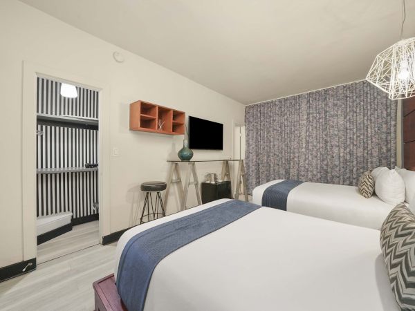 A modern hotel room with two queen beds, blue-gray bed runners, patterned headboard wall, a wall-mounted TV, small desk and stools, and a striped accent wall near the entry.