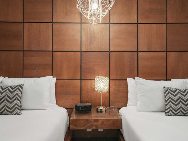 Two neatly made twin beds with white linens and chevron pillows, a shared nightstand, warm lamps, wood-paneled headboard wall, and a geometric chandelier.