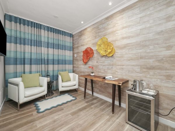 A modern hotel-like sitting area with two white armchairs, green cushions, a striped blue curtain, a small table, wall decor, and a mini fridge with a kettle, all on light wood flooring.