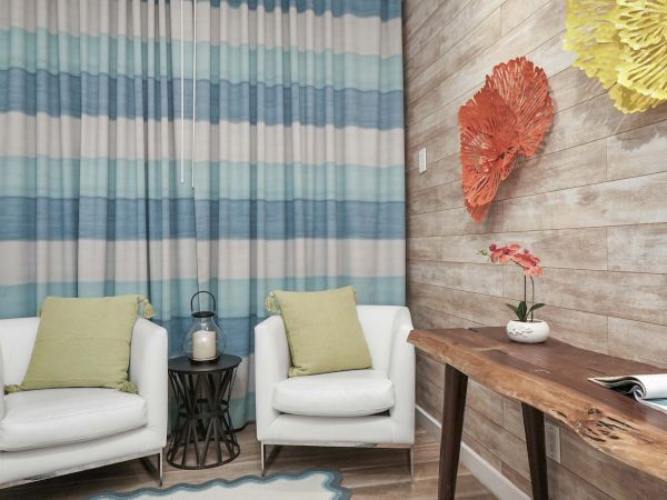 A cozy living corner with two white armchairs, green cushions, a small side table, striped blue curtains, and a wooden desk against a textured wall with bright paper flowers.