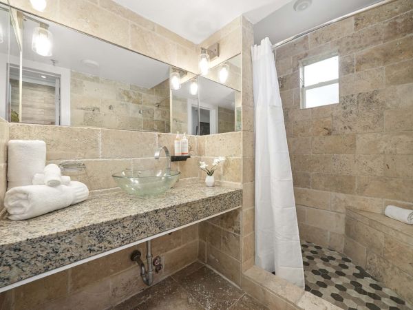 A modern bathroom with beige tiled walls, granite countertop, glass vessel sink, stacked towels, and a separate tiled shower with a small window.
