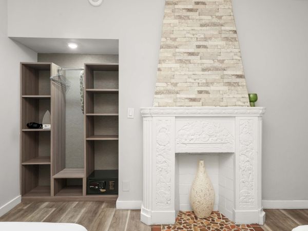 A modern living area with a decorative white fireplace, a brick chimney, and a wooden open shelving unit with a mirror and shoes visible, plus a patterned floor mat, all in neutral tones.