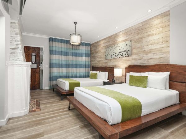 Two neatly made double beds with wooden frames, white linens, green accents, a stone wall, wooden floor, and a seating area in a modern hotel room.