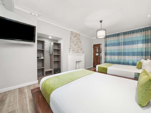 A bright hotel room with two beds, white linens and green accents, a wall of blue striped curtains, a flat-screen TV, and an open closet area near a stone fireplace.