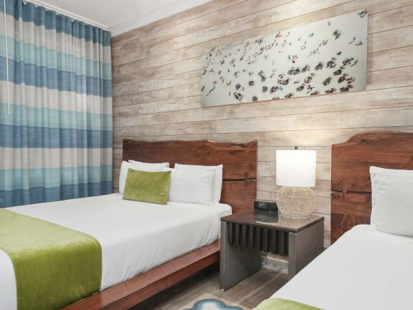 Two hotel beds in a bright room with a nightstand, lamp, and wall art.