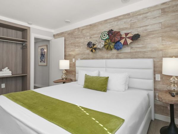 A modern hotel room with a white upholstered bed, green accent throw and pillow, wood-paneled wall, two bedside lamps, and colorful wall art above the headboard.