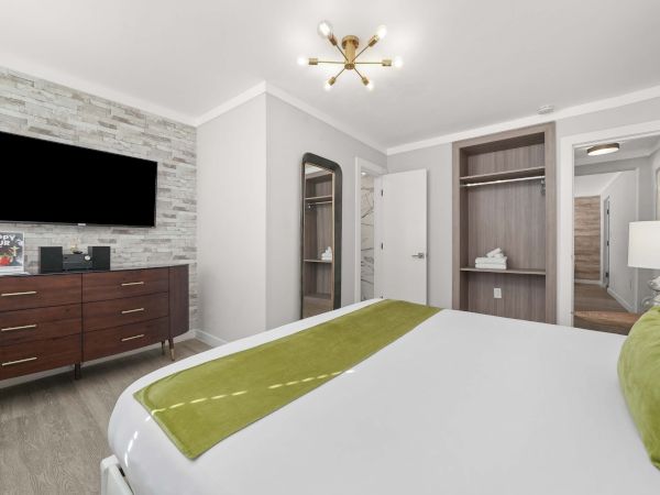 A modern hotel room with a bed, green accent throw, a wall-mounted TV above a wooden dresser, a stone feature wall, and open-wardrobe alcoves.