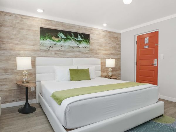 A modern hotel room with a white bed, green accents, two lamps on matching nightstands, stone-inset accent wall, and orange door.