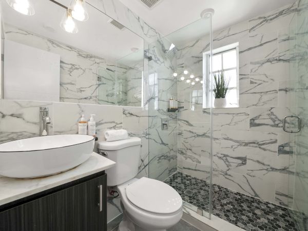 A modern bathroom with marble walls, a glass shower, a white toilet, a vessel sink, and a dark vanity, all in a sleek, bright space.