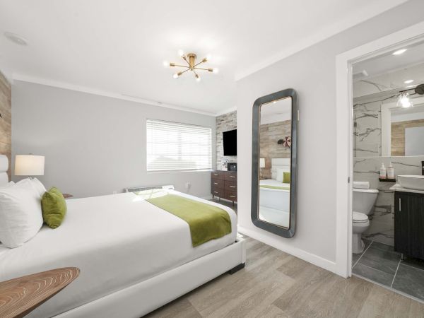 A bright hotel room with a white bed, green accents, a full-length mirror, and an adjacent modern bathroom with marble tiling, all in a clean, airy space.