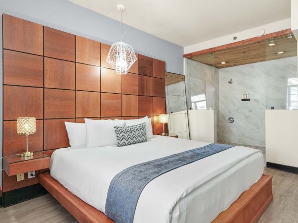 A modern hotel bedroom with a large wooden paneled headboard, white bedding, blue accents, side lamps, and an open glass-walled bathroom.