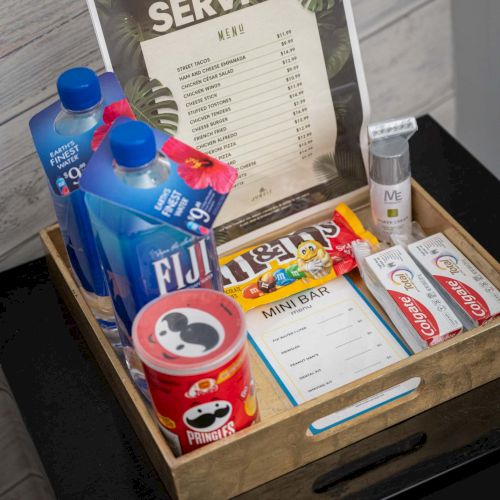 A wooden tray on a black surface holds hotel room amenities: toiletries, snacks, a &ldquo;Room Service&rdquo; menu, coffee creamers, soda, candy, tissues, and a small card. Always ends the sentence.