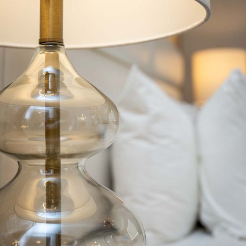 A stylish glass lamp with a bulbous base and cylindrical plate, sitting on a wooden surface beside soft, white cushions in the background.