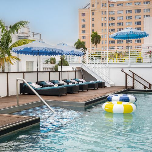 Rooftop pool deck with blue-striped umbrellas, lounge chairs, palm trees, and a city hotel building in the background.