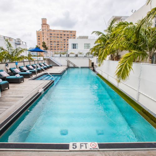 An outdoor rooftop pool with turquoise water, lounge chairs, palm trees, and city buildings under a cloudy sky.