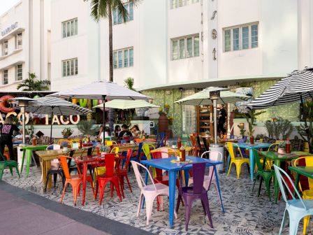 A lively outdoor cafe with colorful plastic chairs and tables under umbrellas, in front of a white building with palm trees and a &ldquo;TACO TACO&rdquo; sign.