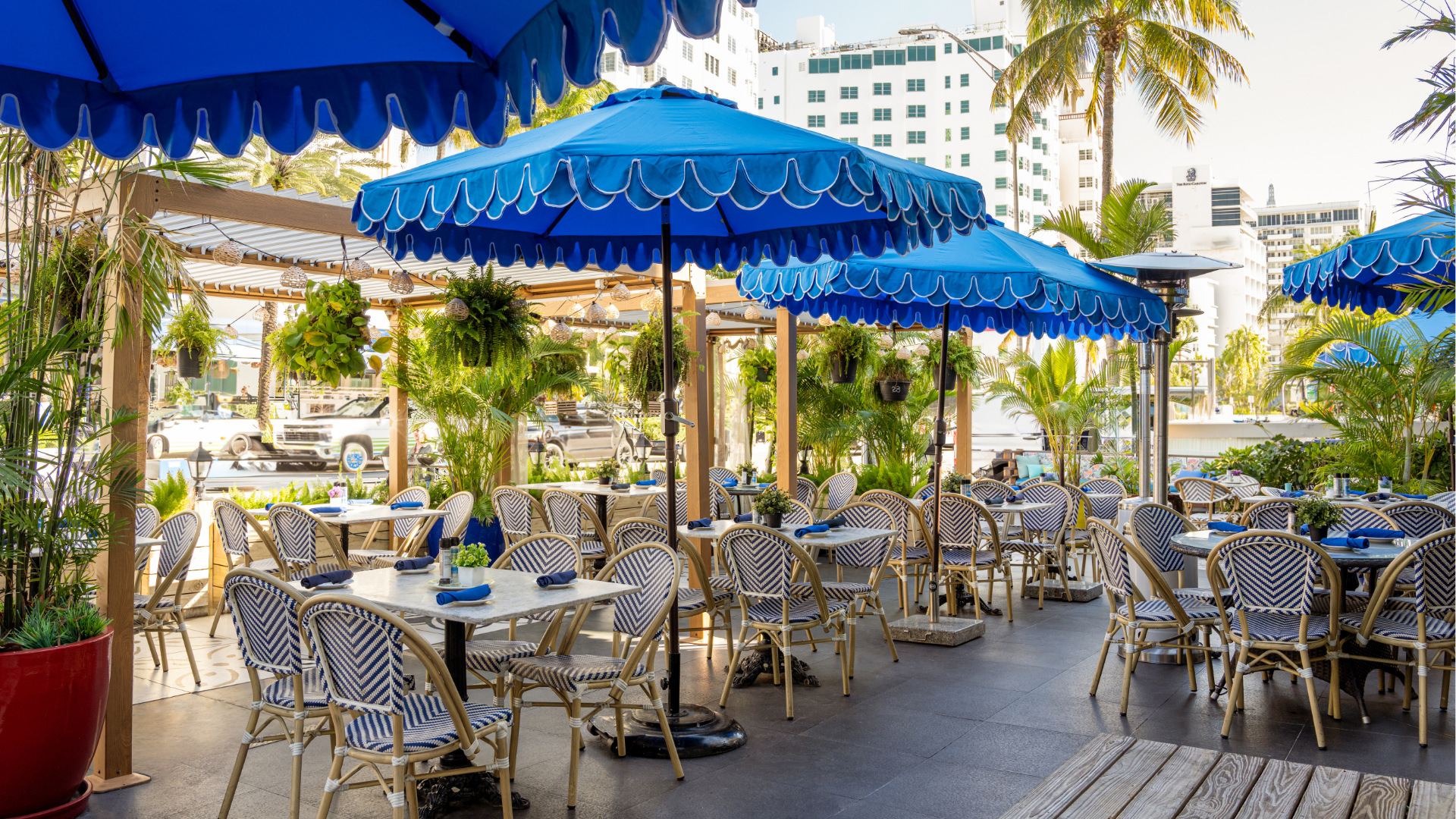 An outdoor bar area with blue umbrellas, tables, and chairs, surrounded by palm trees and a city skyline.