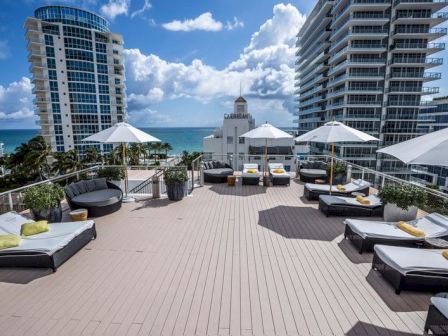 Rooftop pool area with several lounge chairs, white umbrellas, and sunbeds facing the ocean; tall modern buildings nearby under a blue sky.
